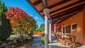 A covered outdoor patio with stone flooring, wooden furniture, and a view of a garden with a tree and a stone pathway.