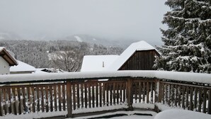 A wooden balcony with snow-covered railings and a table, overlooking a snowy landscape with buildings and trees. - Apartment Margit by Interhome (Haus)