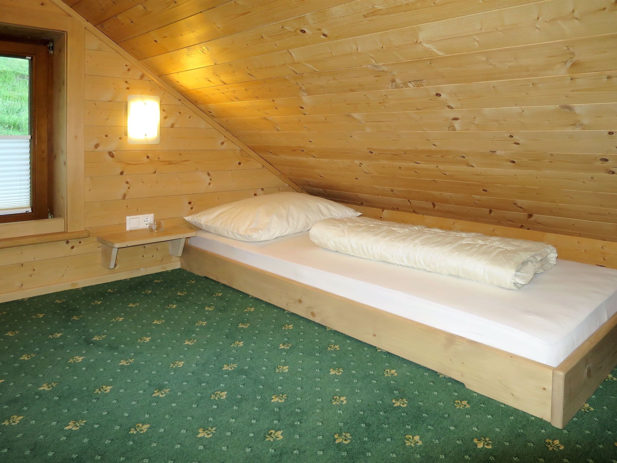 A small, single-bed room with a wooden ceiling and walls, a green patterned carpet, and a window with blinds.