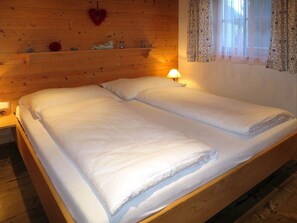 Ein Doppelbett mit weißer Bettwäsche in einem Raum, dessen Wände aus Holz sind, mit einer Herzdekoration und einem Fenster mit gemusterten Vorhängen.