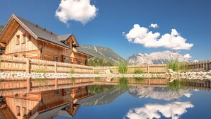 Una casa de madera con balcón, reflejada en un lago tranquilo, rodeada de montañas y un cielo despejado.