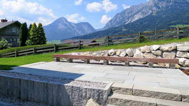 A mountain landscape with a wooden fence, a stone bench, and a house with solar panels.