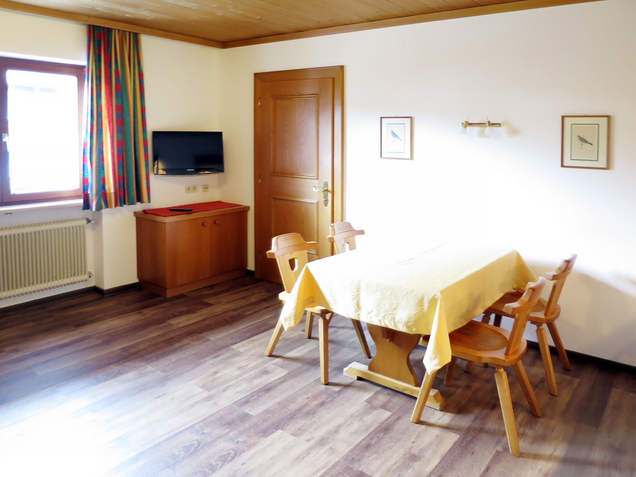 Un comedor con mesa y sillas de madera, un televisor en la pared y una puerta con ventana.