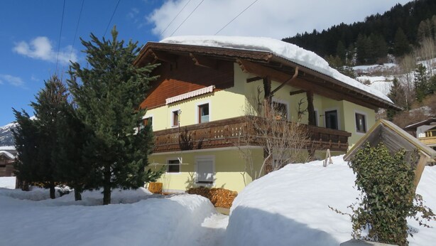 Eine zweigeschossige Chalet mit einem schneebedeckten, geneigten Dach, eingebettet in eine verschneite Landschaft mit Bäumen.