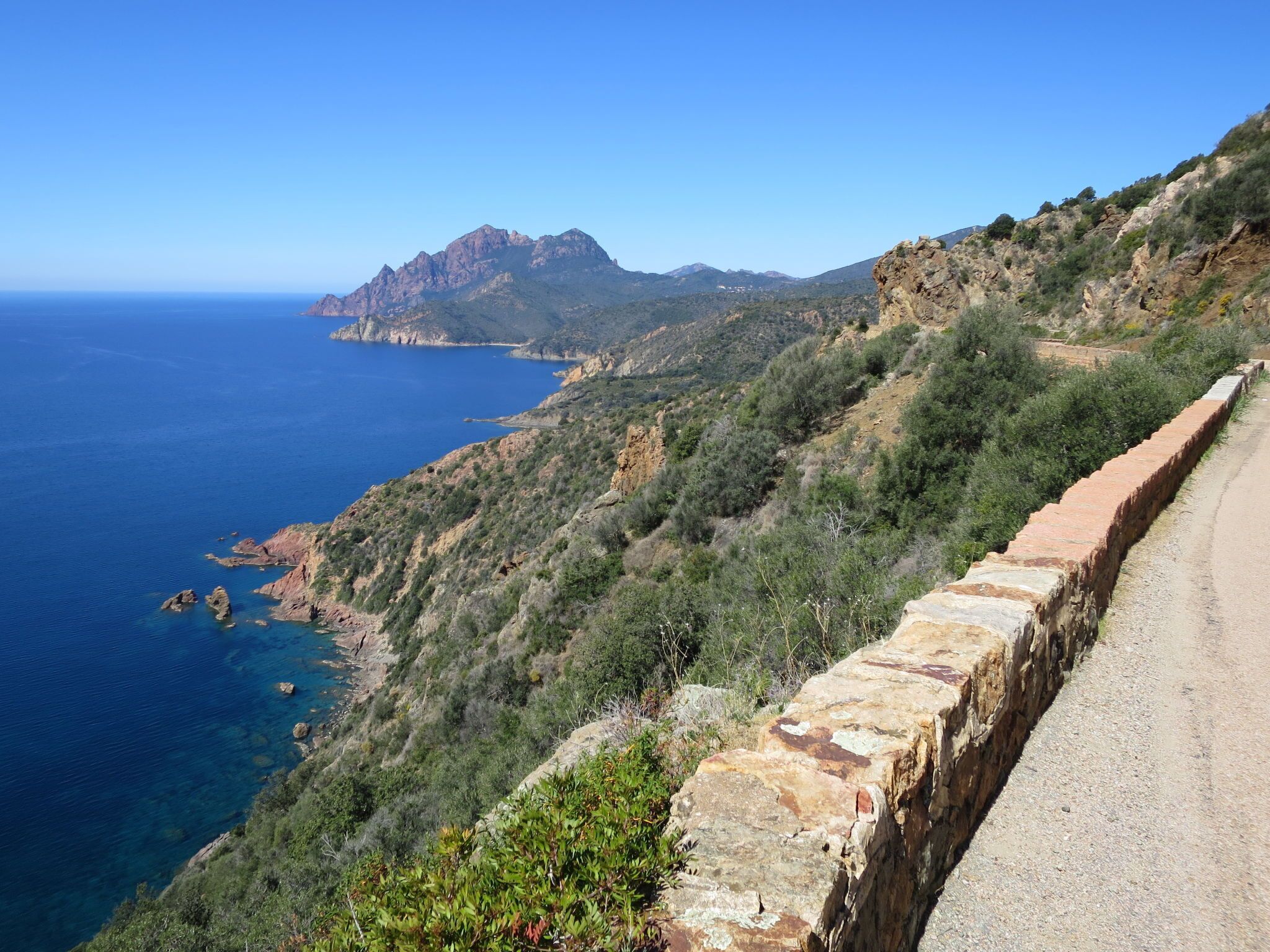 Une route côtière bordée d’un côté par un mur de pierres, offrant de l’autre une vue sur la mer et les montagnes au loin.