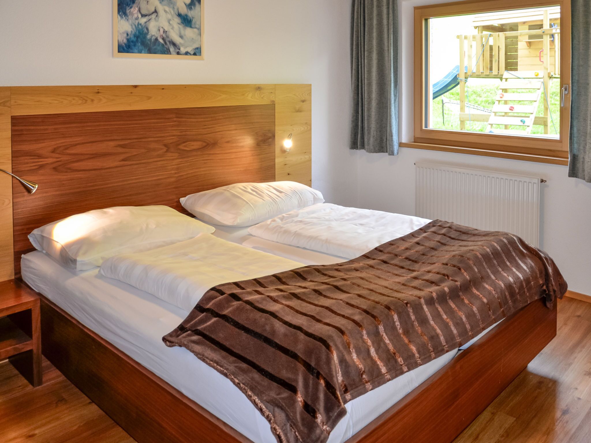 A bedroom with a wooden headboard, white bedding, and a view of an outdoor play area through a window.