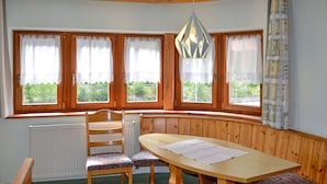 A dining area with a wooden bench, a table, and chairs. There are three windows with curtains, a radiator, and a hanging light fixture.