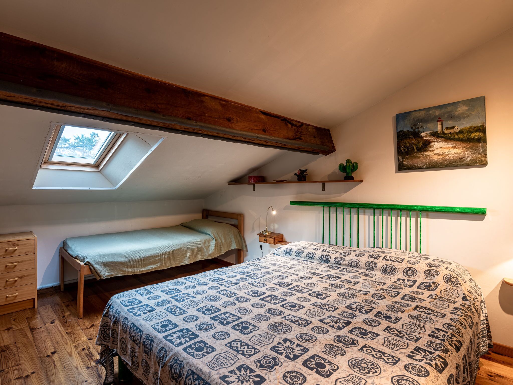 Una camera da letto con soffitto a falde, una finestra a lucernario, un letto con biancheria a motivi, un comodino, una lampada e uno scaffale con un cactus, oltre a un quadro appeso al muro.
