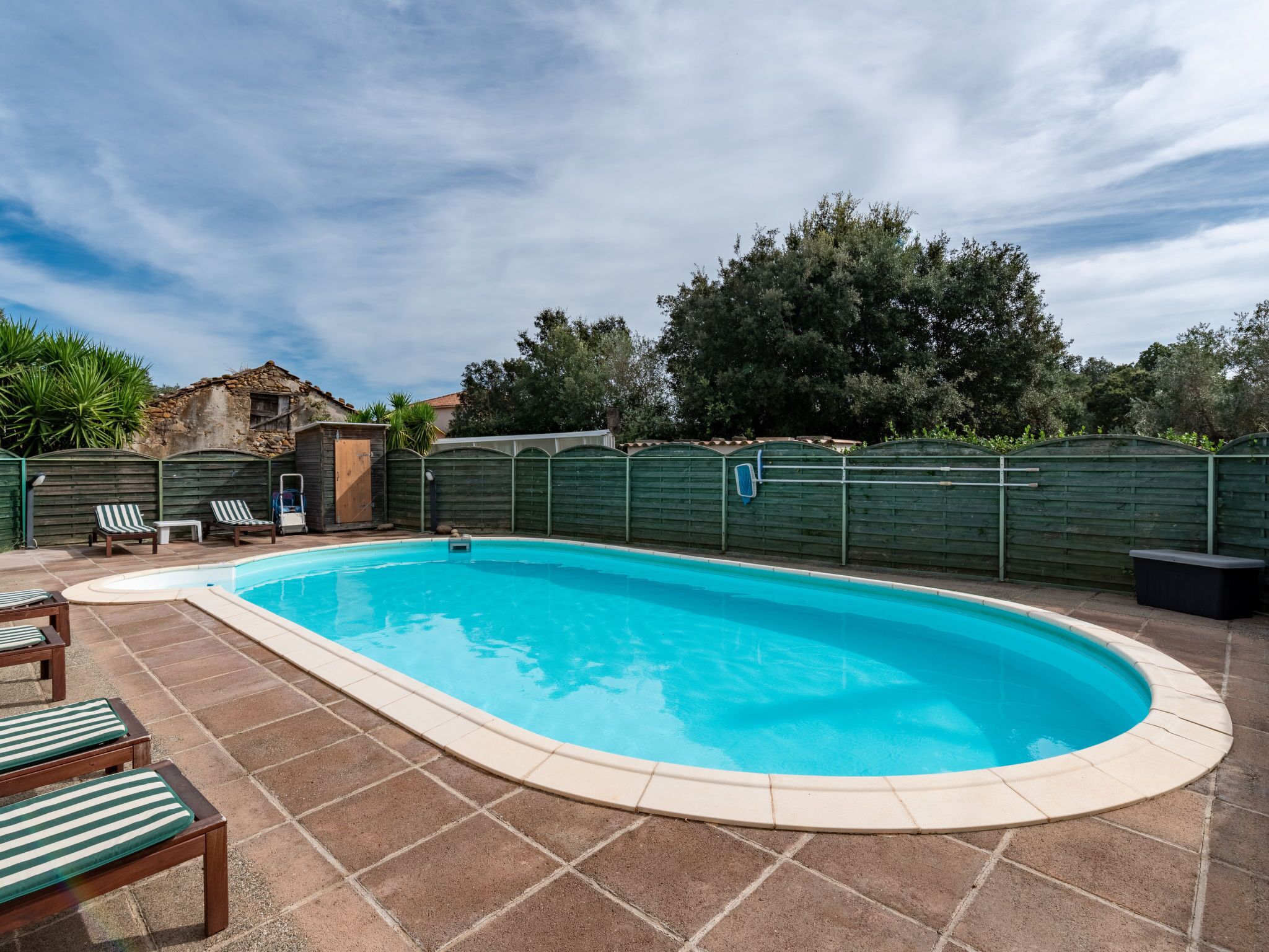 A rectangular swimming pool with a tiled deck, surrounded by a wooden fence and greenery.