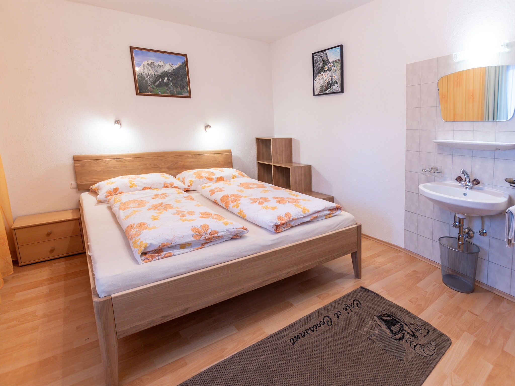 A bedroom with a wooden bed, two wall-mounted lights, a small wooden nightstand, a bedside table, a bathroom with a sink and mirror, and two framed pictures on the wall.