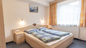 Habitación individual con una cama de madera, mesita de noche, cajón, lámpara de pared, ventana con cortinas y un cuadro en la pared.