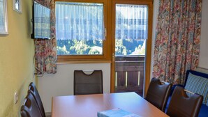 Un coin repas avec une table en bois, quatre chaises, un livre posé sur la table et une vue sur un balcon doté d’une balustrade en bois.