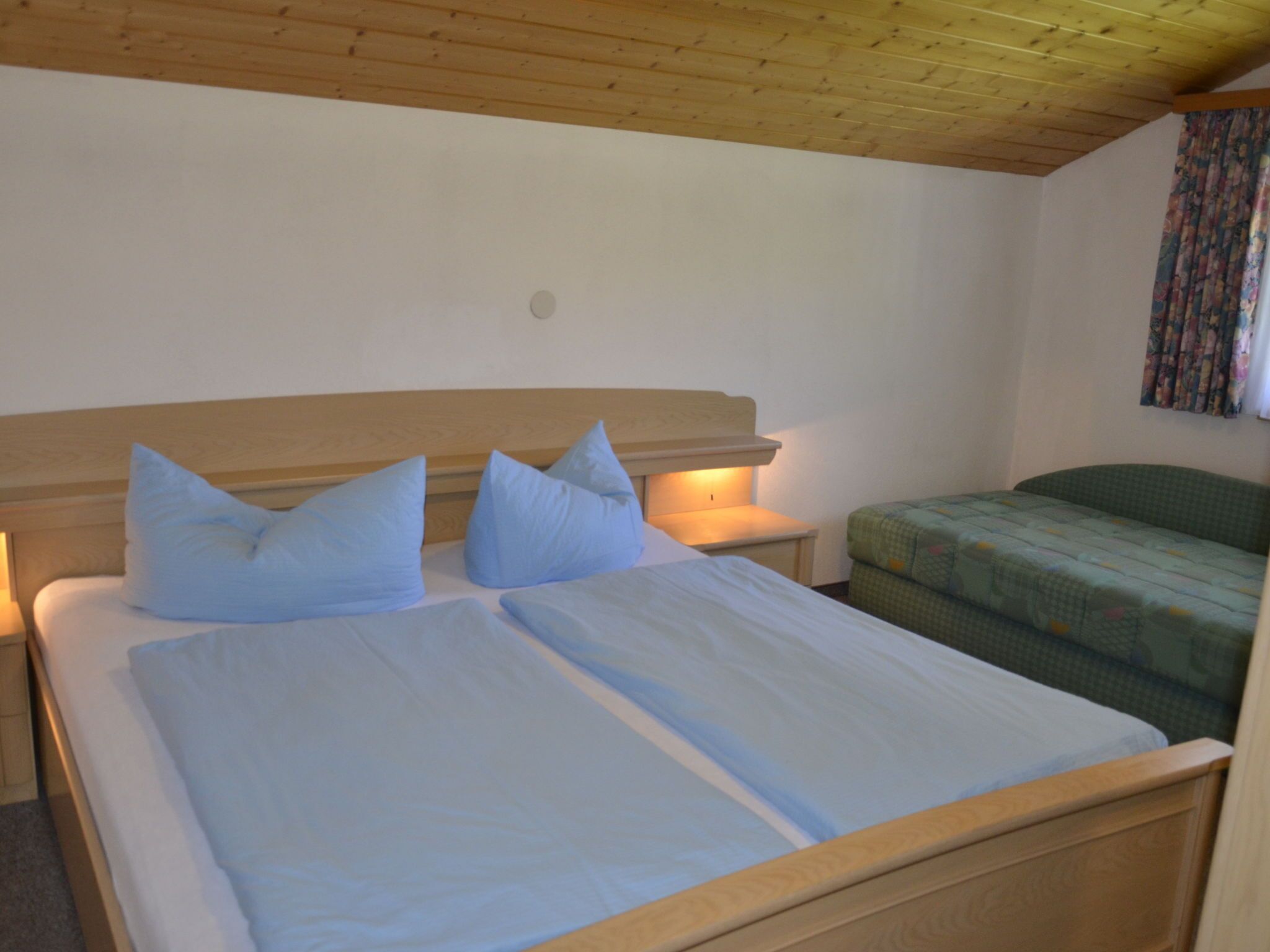 A bedroom with a wooden headboard, two single beds with blue pillows, a green sofa, and a nightstand with a lamp.