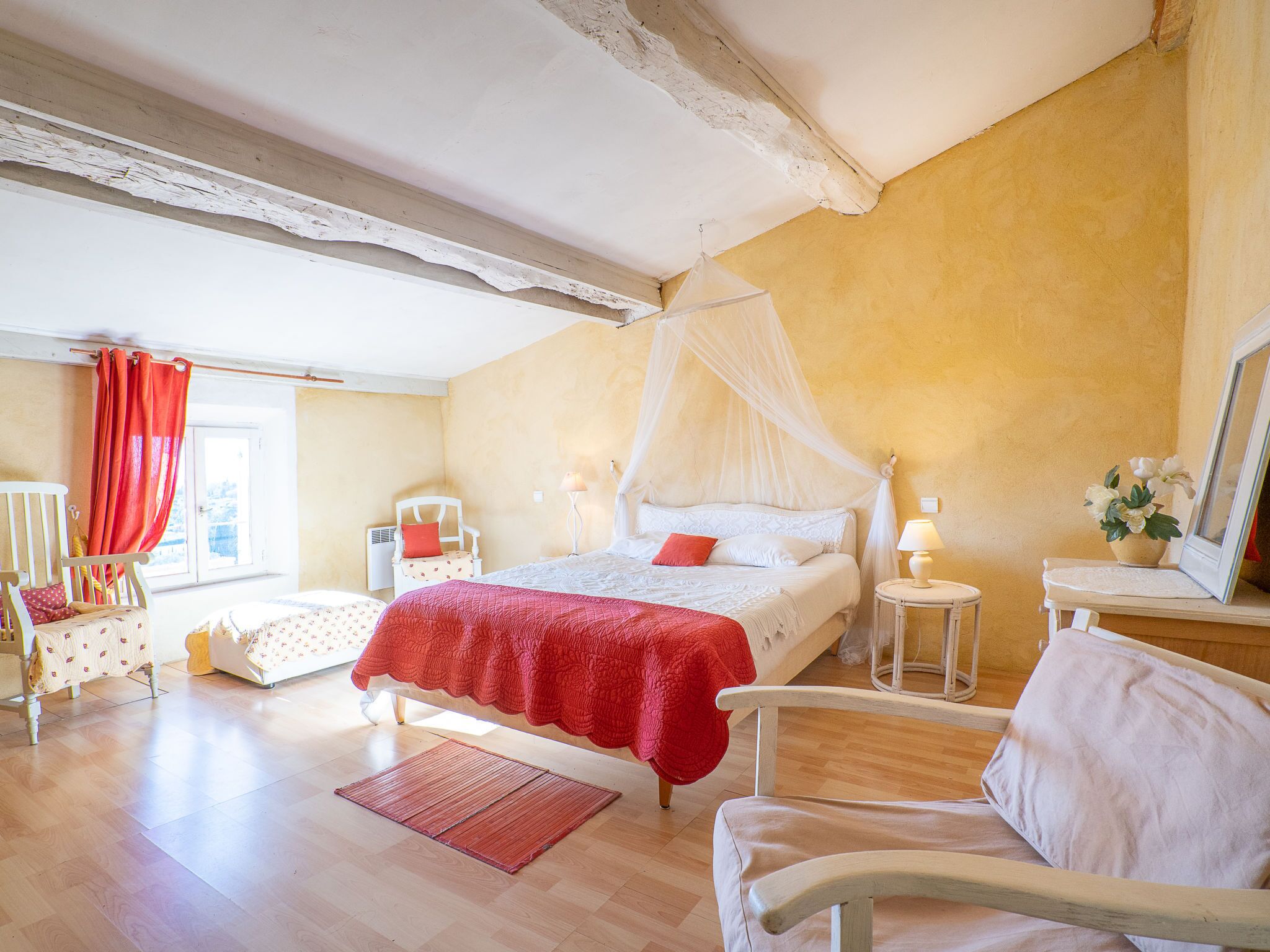 Ein Schlafzimmer mit Bett, Sessel, Nachttisch, Lampe und einem Fenster mit Vorhängen.