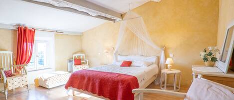 Ein Schlafzimmer mit Bett, Sessel, Nachttisch, Lampe und einem Fenster mit Vorhängen.