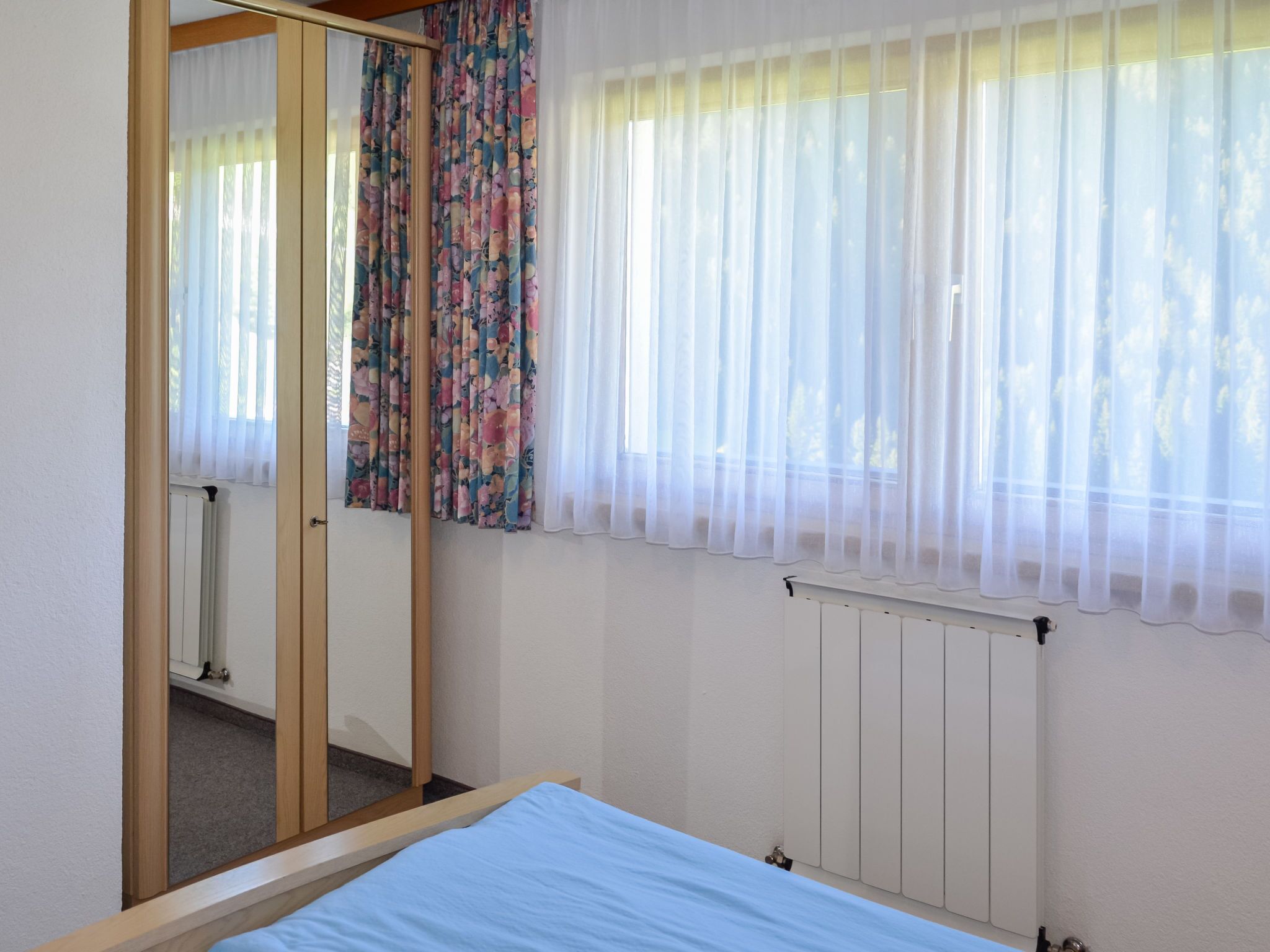 Een slaapkamer met een bed, een spiegel, een radiator en een raam met dunne gordijnen.