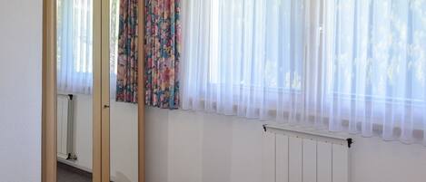 Een slaapkamer met een bed, een spiegel, een radiator en een raam met dunne gordijnen.
