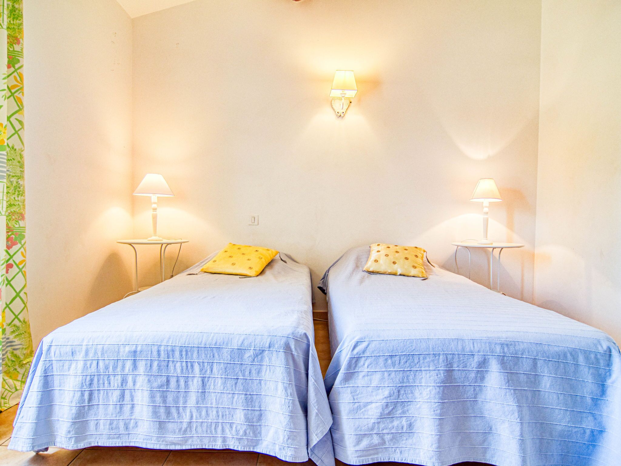 Dos camas individuales con ropa de cama azul a juego y almohadas amarillas, cada una con una mesita de noche y una lámpara de pared.