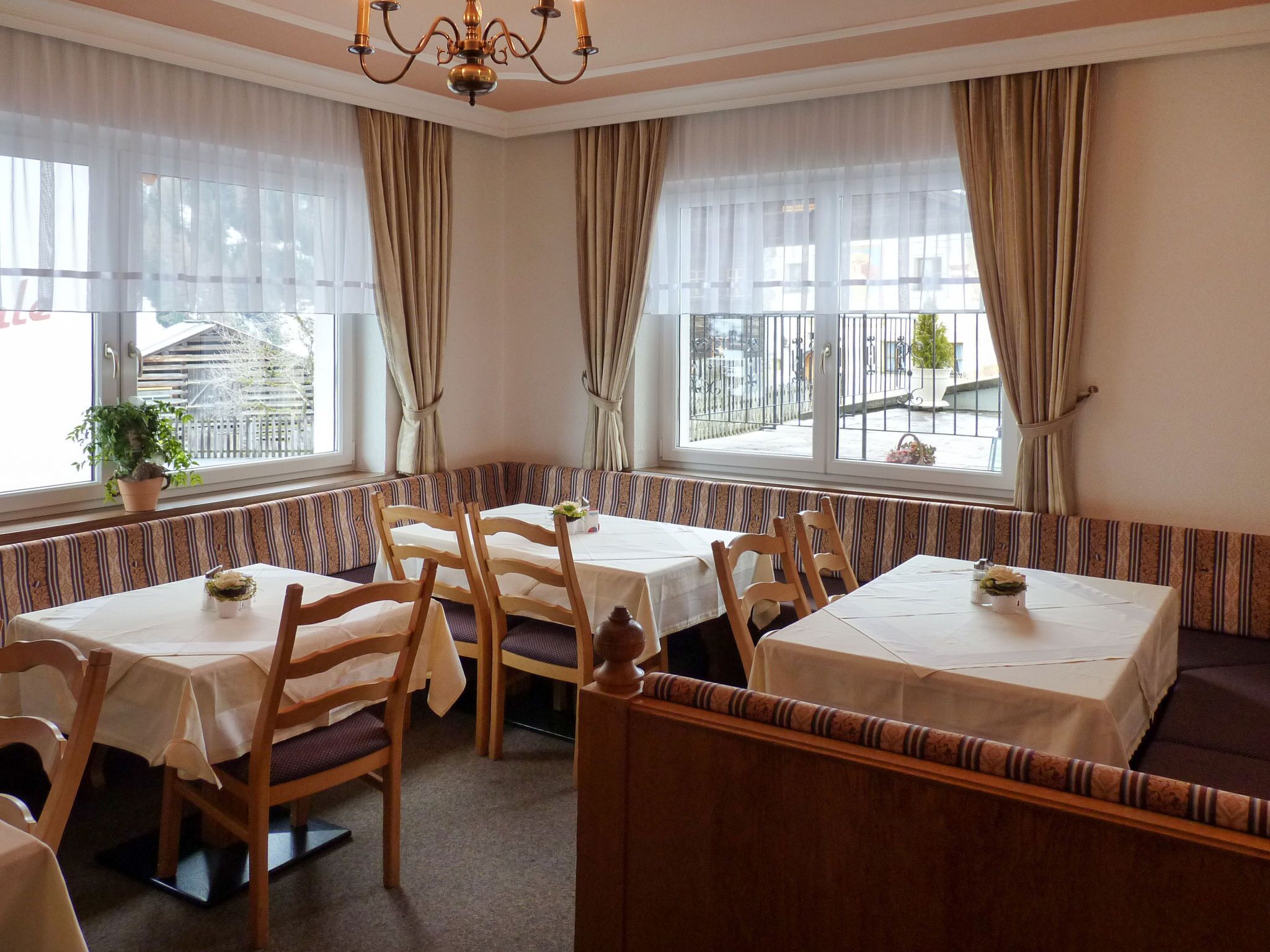 A dining area with tables and benches, large windows with curtains, and a chandelier.