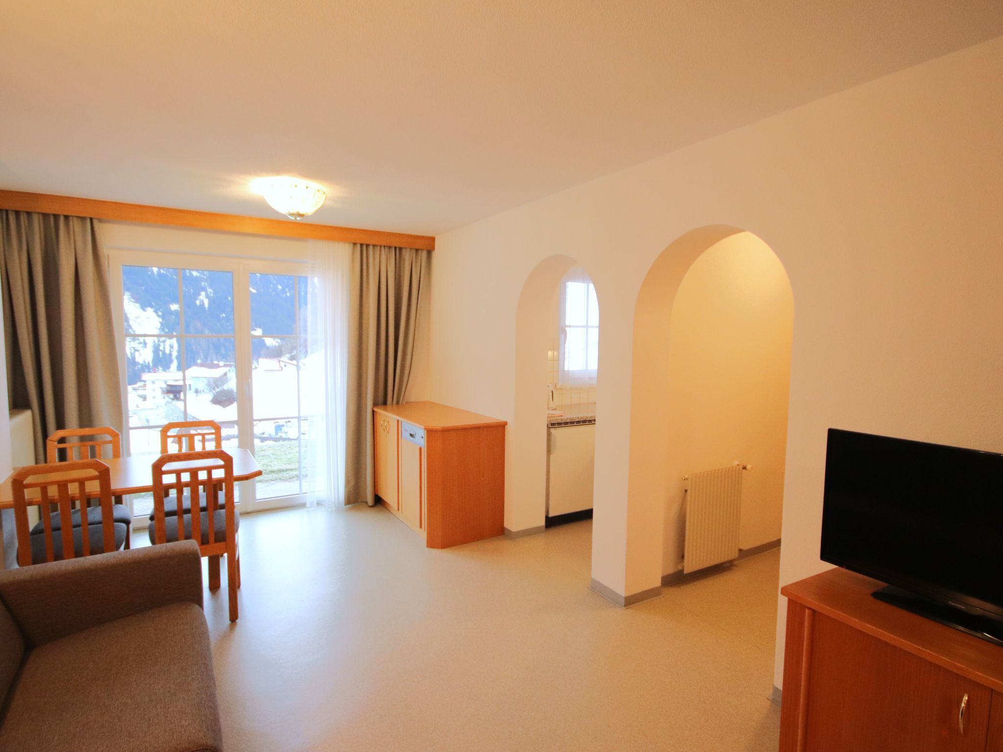 Ein Wohnzimmer mit Sofa, Sesseln, einem Fernseher auf einem Holzkabinett, einer kleinen Küchenzeile und Blick durch ein Fenster.
