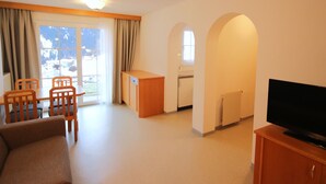 Una sala de estar con sofá, sillones, un televisor sobre un mueble de madera, una pequeña kitchenette y vistas al exterior.