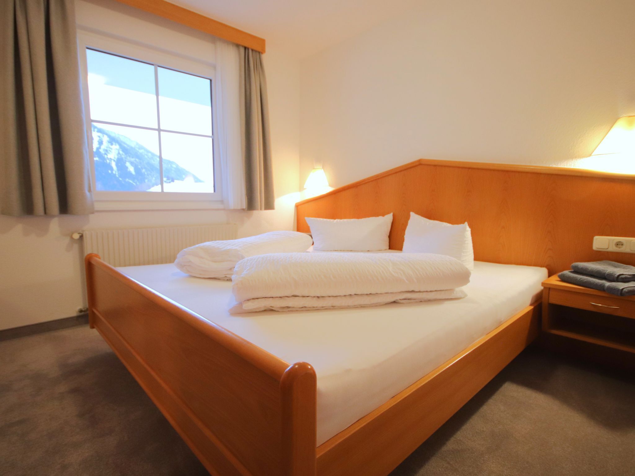 Ein Hotelzimmer mit einem hölzernen Bett, weißen Leinen, einem Nachttisch mit Lampe und einem Fenster mit Vorhängen.