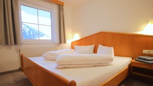 Habitación de hotel con una cama de madera, ropa de cama blanca, una mesita de noche con lámpara y una ventana con cortinas.