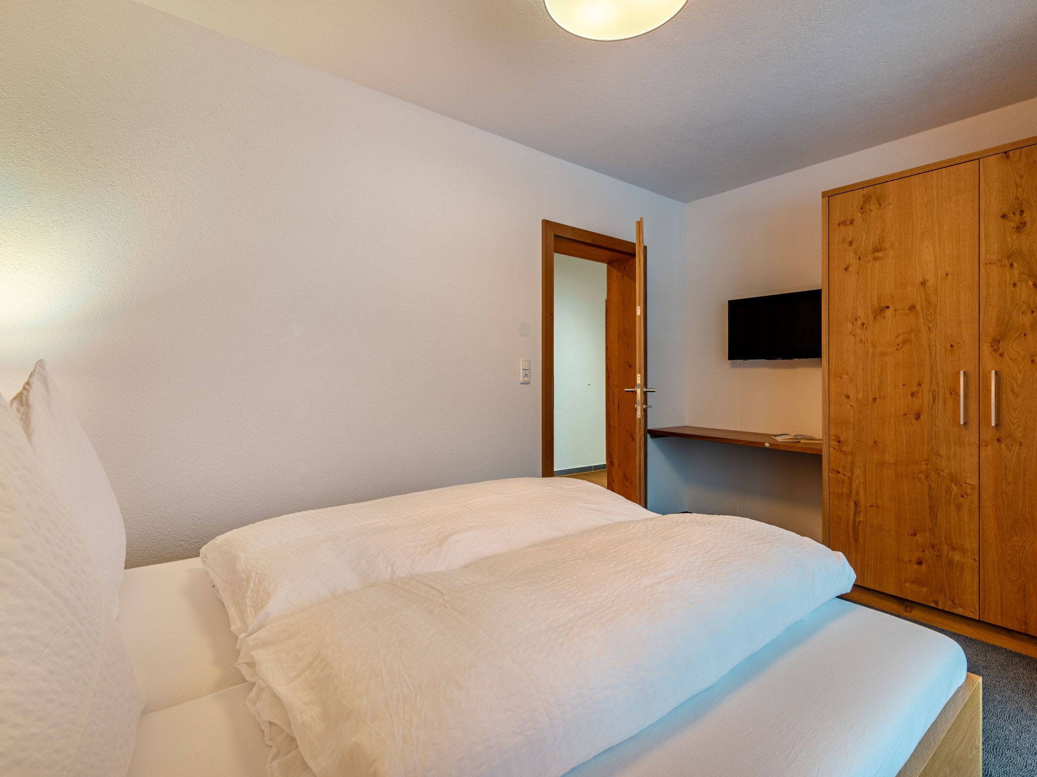 A bedroom with a bed, a wooden wardrobe, and a television on the wall.