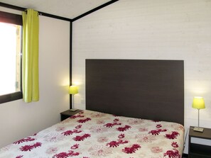 Ein Einzelbett mit floralem Überwurf, zwei Nachttischlampen, ein Fenster mit grüner Vorhang und eine schlichte Wand.
