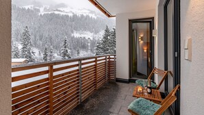 A balcony with a wooden railing, a wooden chair with a furry cushion, and a table with a candle and a bottle.