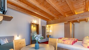 Ein gemütliches Zimmer mit Holzboden, einem runden Holztisch mit Stühlen, einem Bett mit gestreifter Bettwäsche und einem durchgehenden Türspalte sichtbaren Badezimmer.