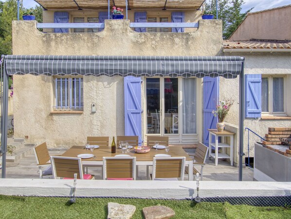A covered outdoor dining area with a table set for four, surrounded by chairs, and a view of a building with blue shutters and a balcony.
