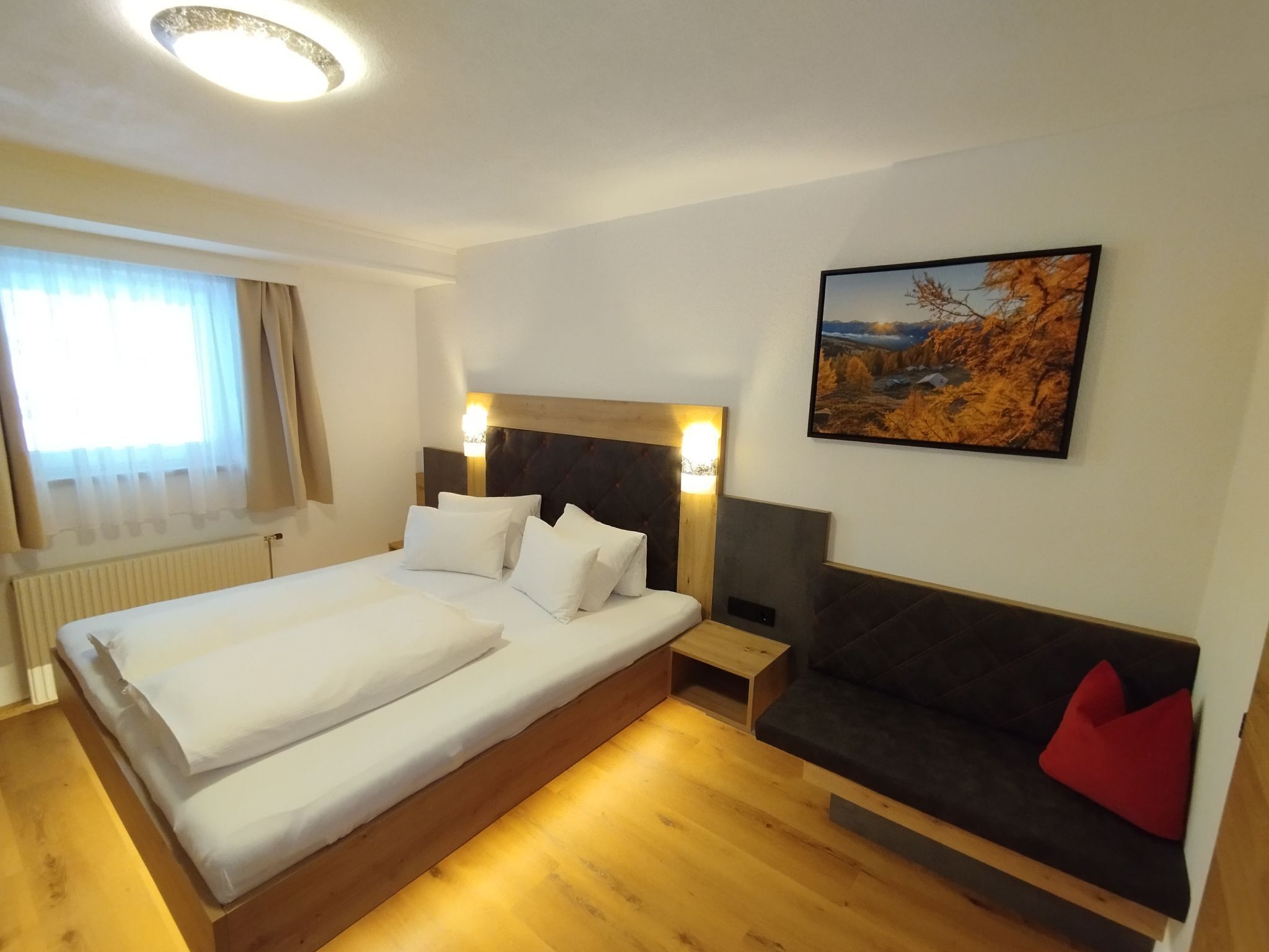 Una camera d'albergo con un letto grande, pavimento in legno, un divano scuro con un cuscino rosso, un quadro appeso al muro e una finestra con tende.