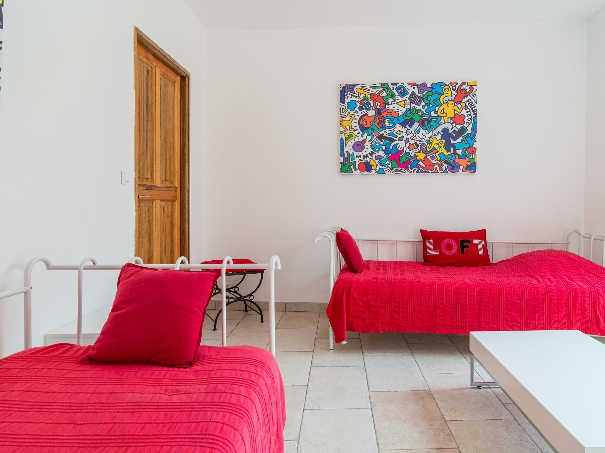 Une chambre avec deux lits, chacun recouvert d’une courtepointe rouge assortie à des coussins, une table blanche et une peinture abstraite aux couleurs vives accrochée au mur.