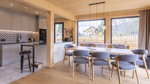 A modern kitchen with wooden cabinets, a dining area with a large table and chairs, and a view of a wooden house through large windows. - #7b mit Sauna & Pool by Interhome (Sankt Georgen am Kreischberg)