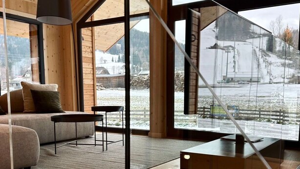Ein modernes Interieur mit Holzbalkendecke, großen Fenstern, einer Couch, einem Tisch und einer Pendelleuchte.