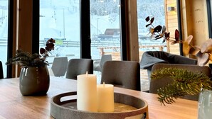 Ein Holzinterieur mit einem Esstisch, Kerzen und Blick auf eine verschneite Landschaft durch große Fenster.