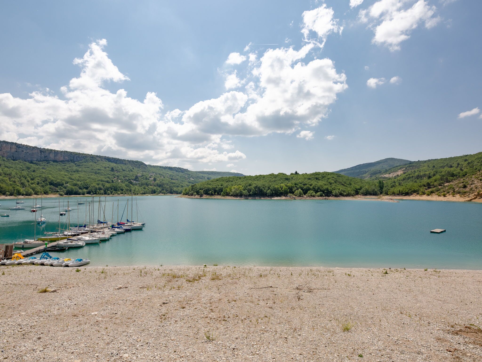 Un lago tranquilo con barcos amarrados en la orilla, rodeado de colinas y cielos despejados.