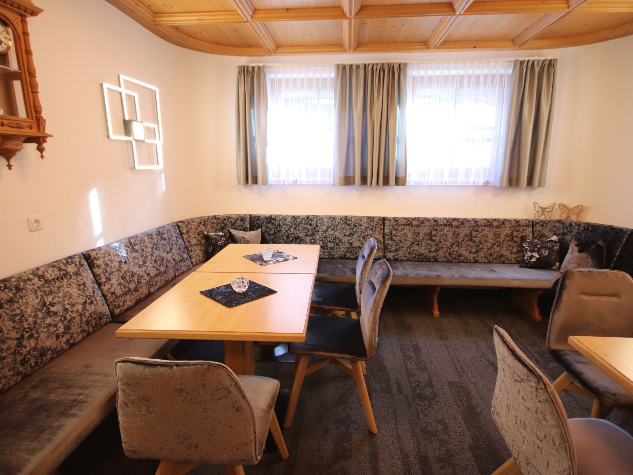 A room with a wooden ceiling, a large window with curtains, a wooden table, and a seating area with a bench and chairs.