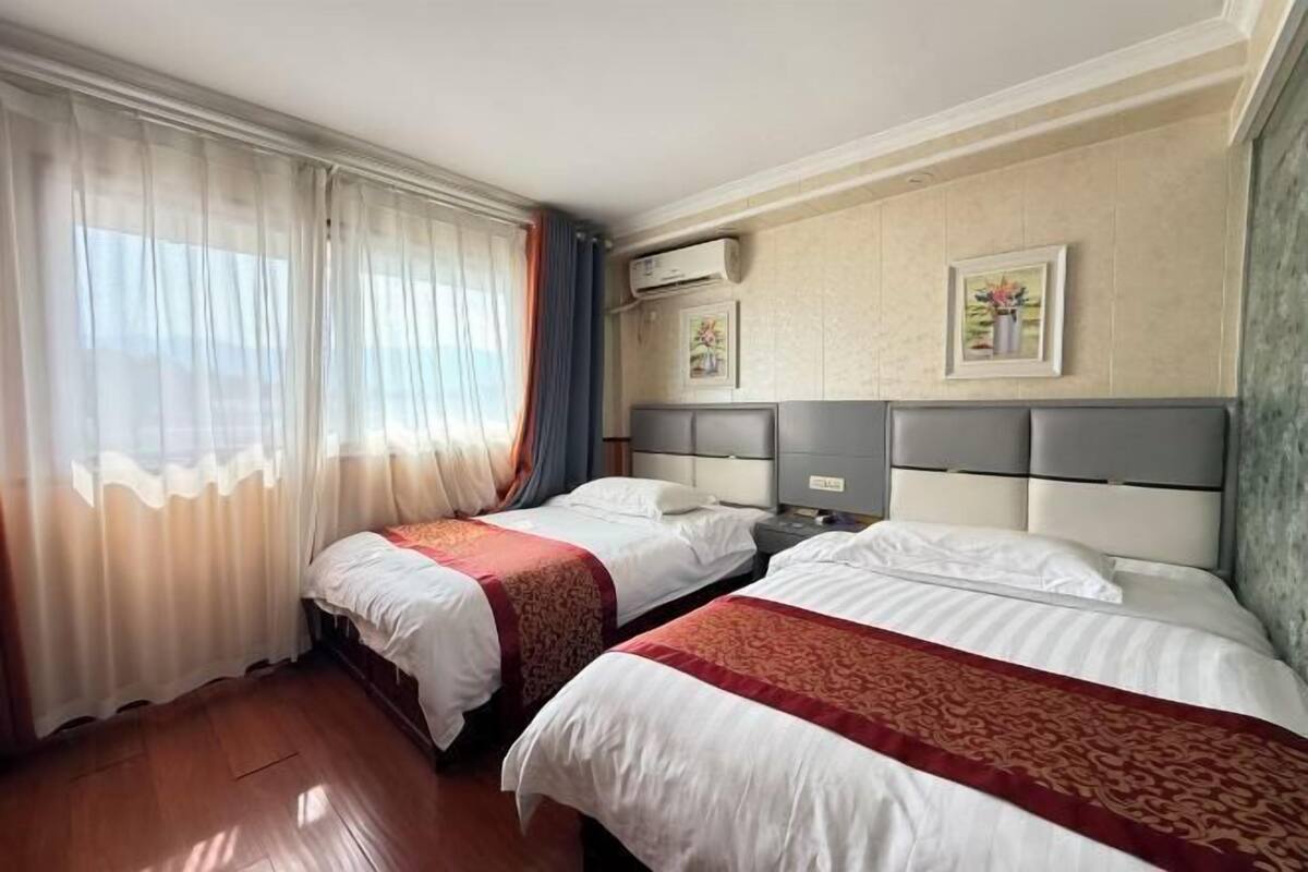 Habitación de hotel con dos camas, una ventana con cortinas traslúcidas, un equipo de aire acondicionado y dos cuadros en la pared.