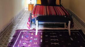 Un couloir étroit, avec un lit au fond, un petit tabouret en bois et un tapis à motifs sur le sol.