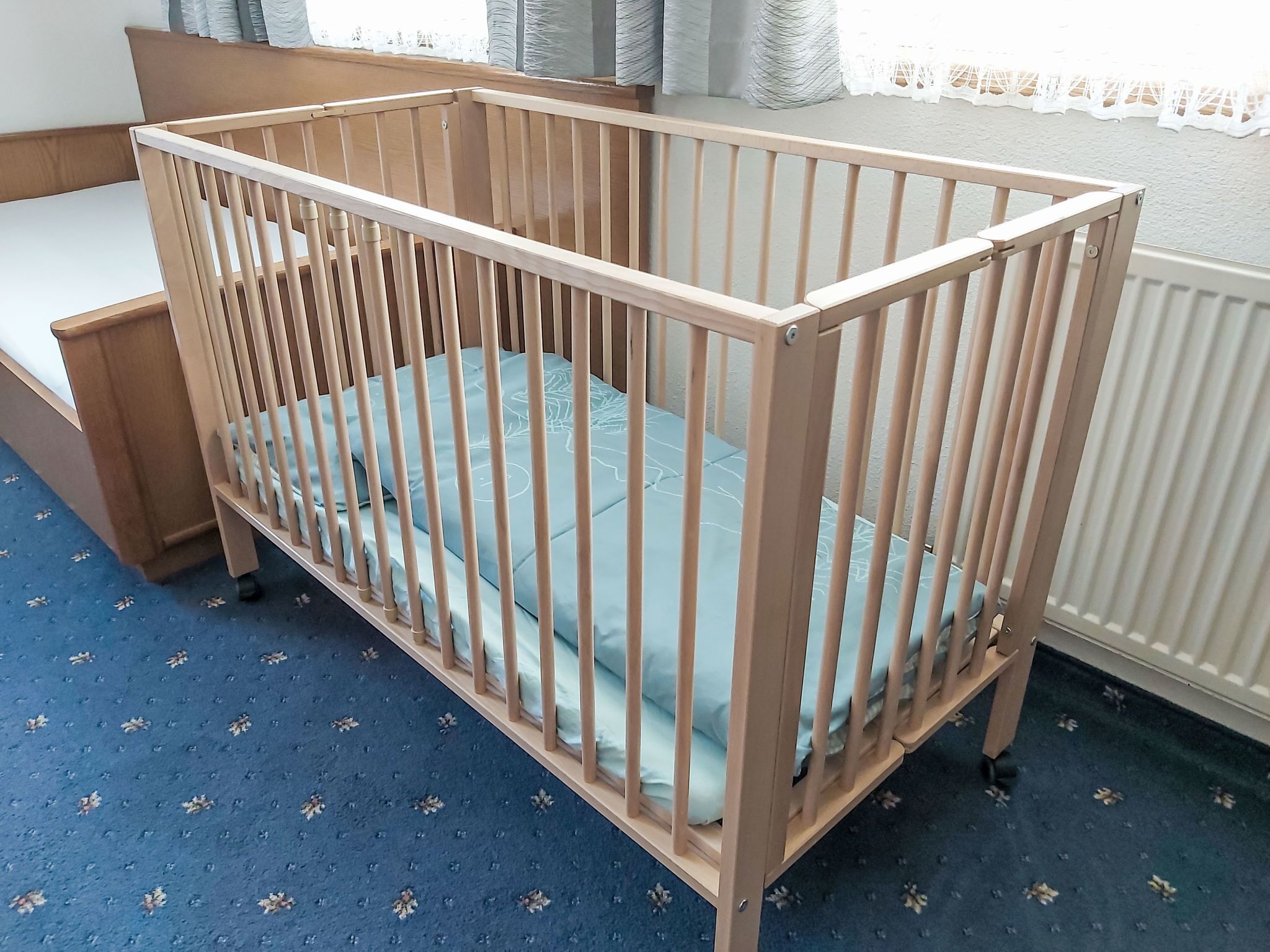 Ein Holzkindbett mit einer hellblauen Matratze in einem Zimmer mit blauem Teppich und einem weißen Heizkörper.