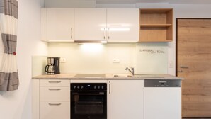 Een kleine keuken met witte kasten, een inbouwoven, een koffiezetapparaat en een houten plank.