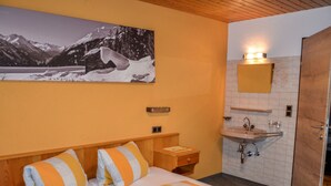 Een hotelkamer met een bed, een nachtkastje, een badkamer met een wastafel en spiegel, en een groot schilderij van een besneeuwd berglandschap aan de muur.