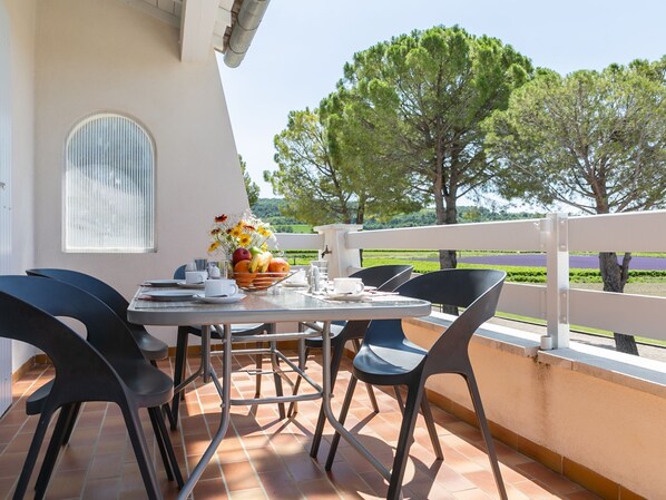 Um terraço com uma mesa posta para refeições, rodeado por cadeiras, uma janela e vista para árvores e um campo.