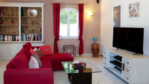 A living room with a red sofa, a glass coffee table, a bookshelf, a flat-screen TV, and a window with red curtains. - Sweet Home in Luberon by Interhome (Villelaure)