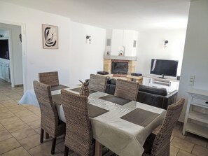 Uma sala de jantar com cadeiras de vime e uma mesa, lareira, televisão e cozinha ao fundo.