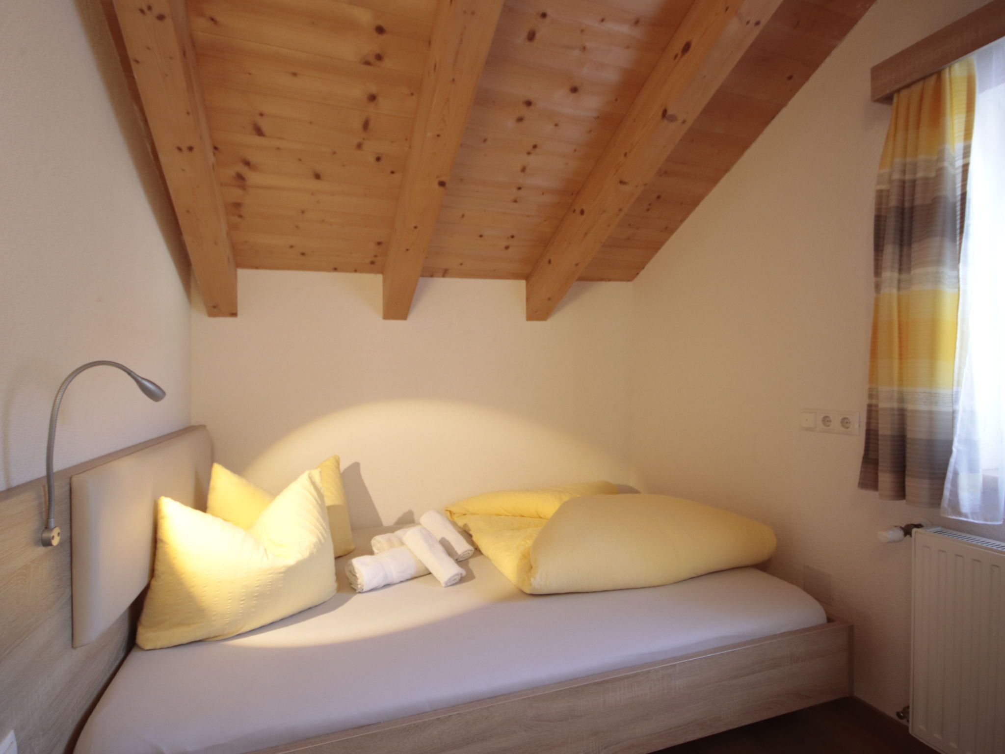 A small, cozy bedroom with a wooden ceiling, a bed with white bedding, and a window with a curtain.