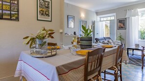 Un coin repas avec une table dressée pour quatre personnes, entourée de chaises en bois. On peut voir deux plantes en pot sur la table, une bouteille de vin et un verre. La pièce a un plancher de couleur claire, et les murs sont ornés de tableaux encadrés.