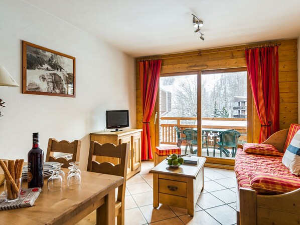 A cozy room with a wooden desk, a television, a dining table, and a sofa. There is a window with curtains and a view of trees outside.
